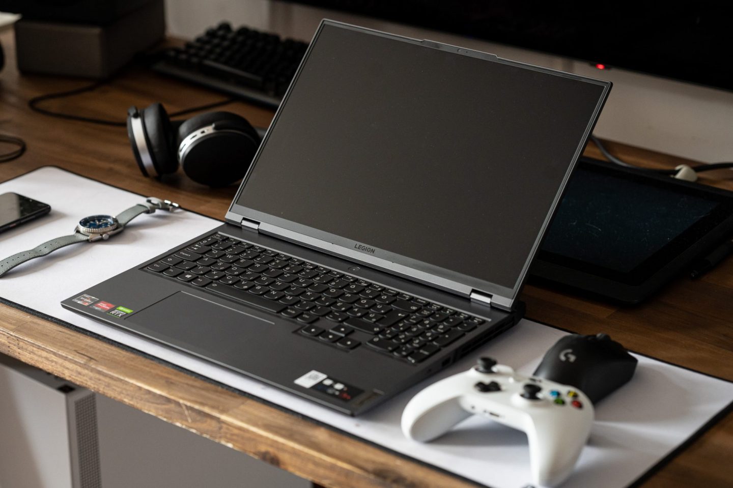 Nyitott Lenovo Legion gamer laptop az asztalon, előtte egy fehér kontrollerrel, fekete egérrel, karórával és fejhallgatóval.