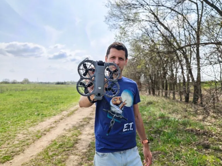 Férfi mutatja a kompaktnak tűnő DJI Neo 2 drónt a szabadban, zöld mező előtt.