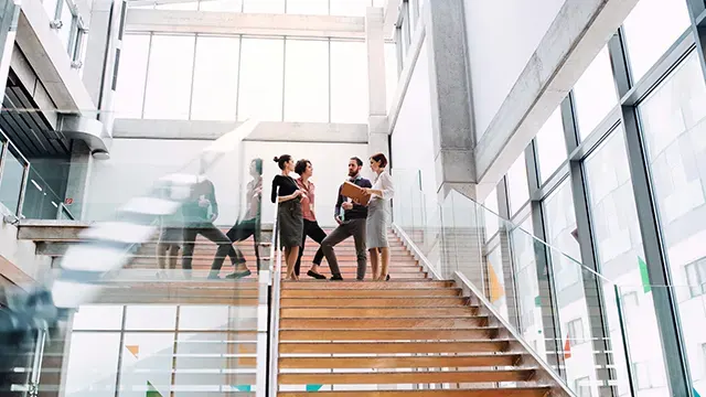 Négy ember beszélget a fa lépcsőn, nagy ablakok és üvegkorlátok mellett egy modern épületben.