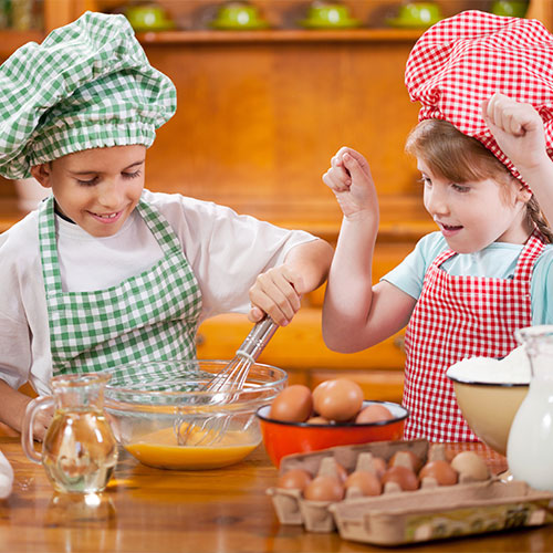 Két gyerek szakácssapkában és kötényben. Egyikük habverővel kever egy tálban, a másikuk ökölbe szorított kézzel nézi.