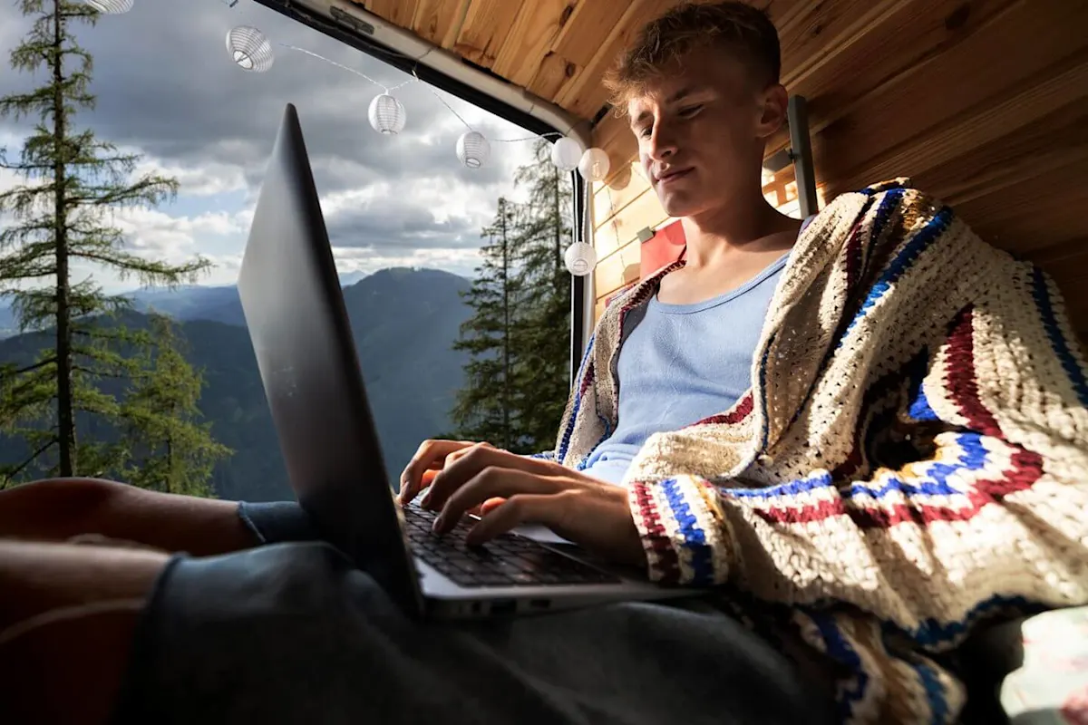 Fiatal férfi laptopon dolgozik egy otthonos lakóautóban, háttérben fenyőerdővel és hegyekkel.
