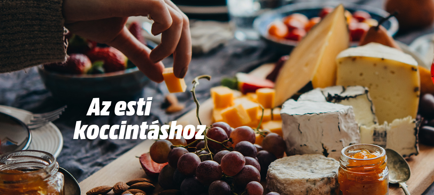 Az esti koccintáshoz felirat, sajtok, szőlő, lekvár és mandula látható egy fa deszkán. Egy kéz egy sajtkockát emel fel.