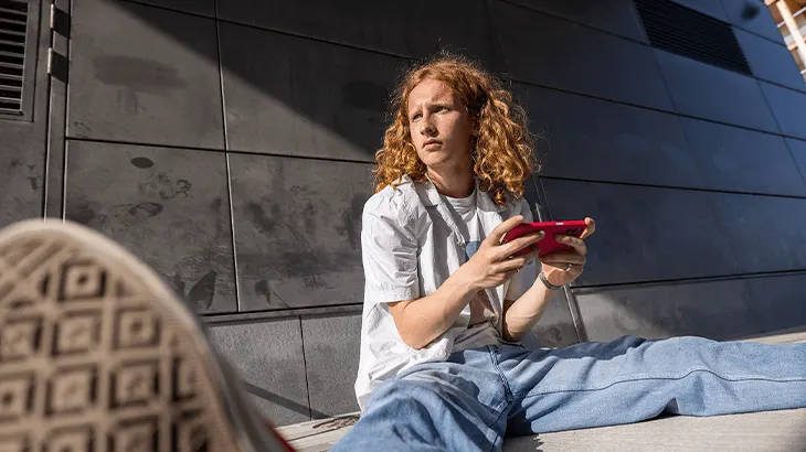 Fiatal ül kék farmerben, fehér pólóban, piros telefont tartva. Egy cipő látható, háttere szürke fal.