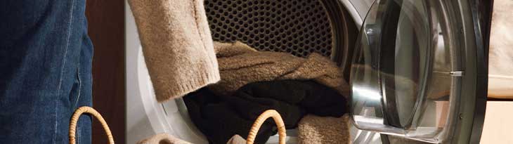 Egy nyitott szárítógépben ruhák vannak, mellette egy farmernadrágot viselő személy kosarat tart.