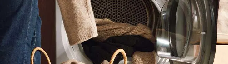 Egy nyitott szárítógépben ruhák vannak, mellette egy farmernadrágot viselő személy kosarat tart.