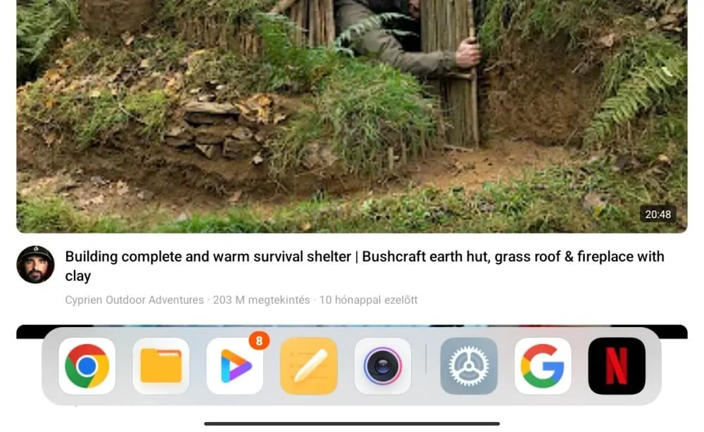 Képernyőkép egy YouTube videóról egy bushcraft menedék építéséről, alul a tableten megnyitott alkalmazások dokkoló sávjával.