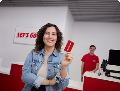 Egy nő farmerdzsekiben ajándékkártyát tart, mosolyog. Háttérben egy férfi piros pólóban áll pult mögött, számítógép látható.