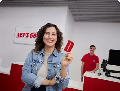 Egy nő farmerdzsekiben ajándékkártyát tart, mosolyog. Háttérben egy férfi piros pólóban áll pult mögött, számítógép látható.