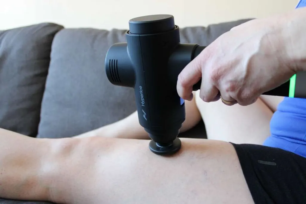 Kék felsőt viselő személy a combizmait kezeli egy fekete Hyperice Hypervolt 2 Pro masszázspisztollyal a kanapén ülve.
