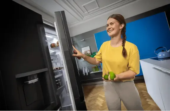 Nő nyitja a hűtőt, zöld almát tartva. A háttérben kék fal és konyhai eszközök láthatóak.