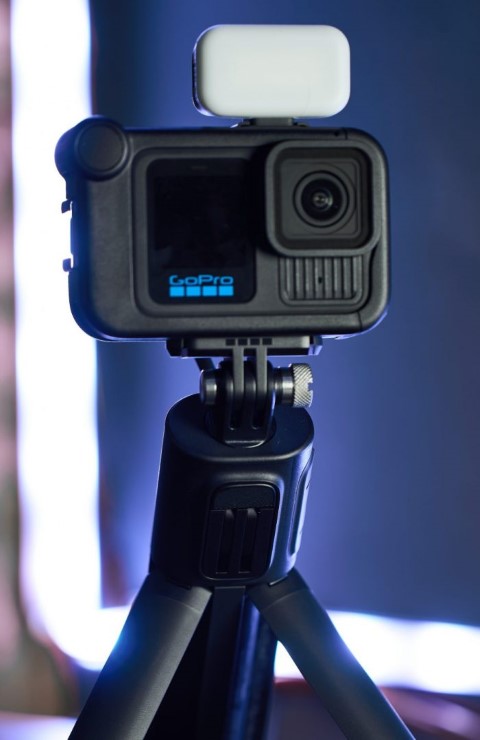 GoPro HERO13 Black akciókamera Media Mod keretben, tetején egy világító fehér LED lámpával, szétnyitott háromlábú állványra (tripodra) rögzítve, kékes háttérvilágítás előtt.