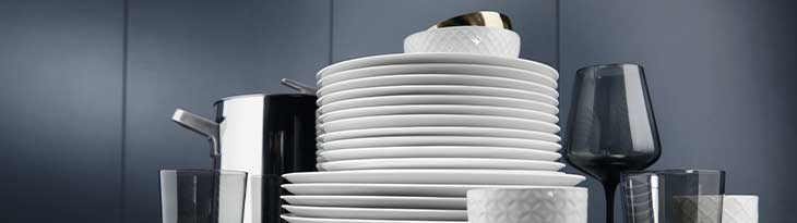 Fehér tányérok, edények, poharak láthatók, részben egymásra helyezve, sötétkék háttér előtt.