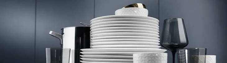 Fehér tányérok, edények, poharak láthatók, részben egymásra helyezve, sötétkék háttér előtt.