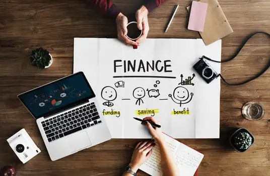 Felülnézeti kép egy fa íróasztalról, rajta laptop, jegyzetek és egy "FINANCE" feliratú rajz a finanszírozási lépésekről.