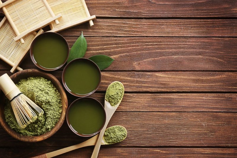 Matcha tea por tálban, bambusz ecset, csészék zöld teával és fakanalak fa asztalon.