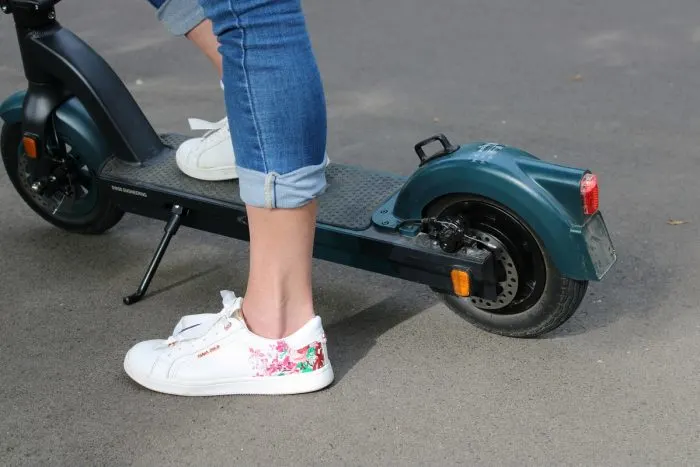 A képen egy sötétkék elektromos roller hátsó része látható, mellette egy fehér, virágmintás tornacipőt és felhajtott szárú farmert viselő személlyel.