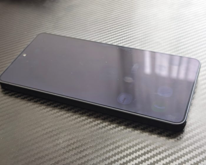 Kikapcsolt kijelzőjű Samsung Galaxy S24 Ultra okostelefon lapos szögben fektetve, karbon mintás felületen.