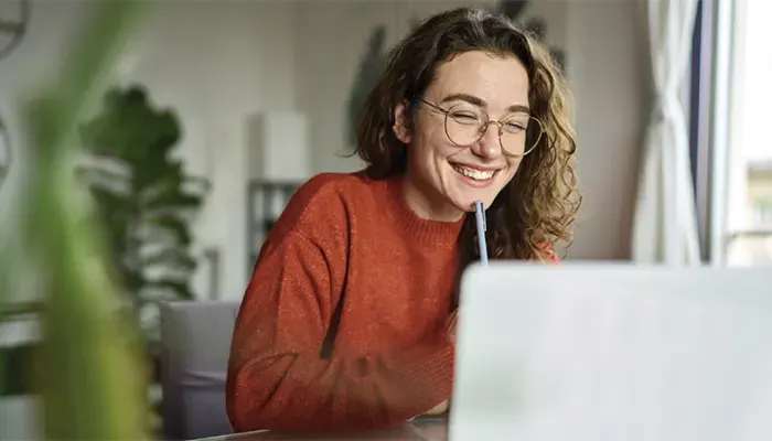 Mosolygó nő narancssárga pulóverben, szemüvegben, tollal az ajka alatt, laptop előtt ülve. Növény és ablak a háttérben.