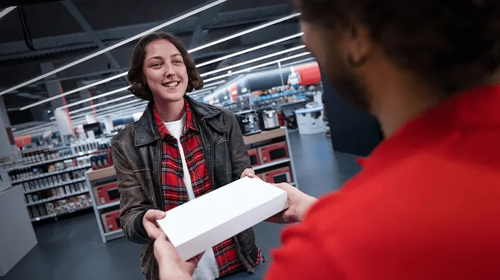 Egy nő kockás inget és kabátot visel, átad egy fehér dobozt egy piros inget viselő személynek egy üzletben.