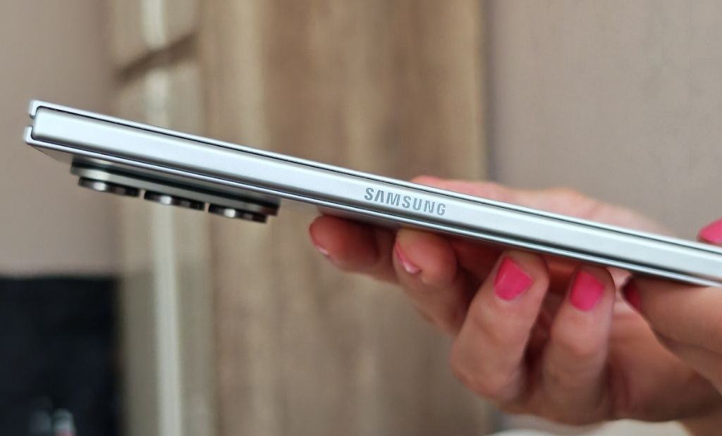 Oldalnézetből fotózott ezüst színű Samsung okostelefon kézben tartva, kiemelkedő kameramodullal.