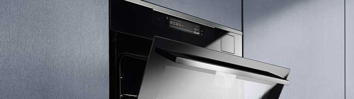 Szürke falba épített, félig nyitott, rozsdamentes acél sütő fekete belsővel és digitális kijelzővel látható.