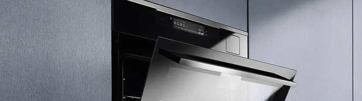 Szürke falba épített, félig nyitott, rozsdamentes acél sütő fekete belsővel és digitális kijelzővel látható.