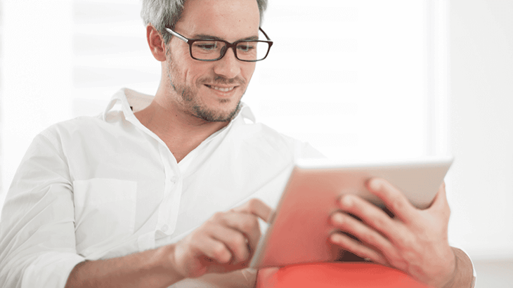 Férfi szemüvegben, fehér ingben, táblagépet használ narancssárga háttér előtt, fehér falak mellett.