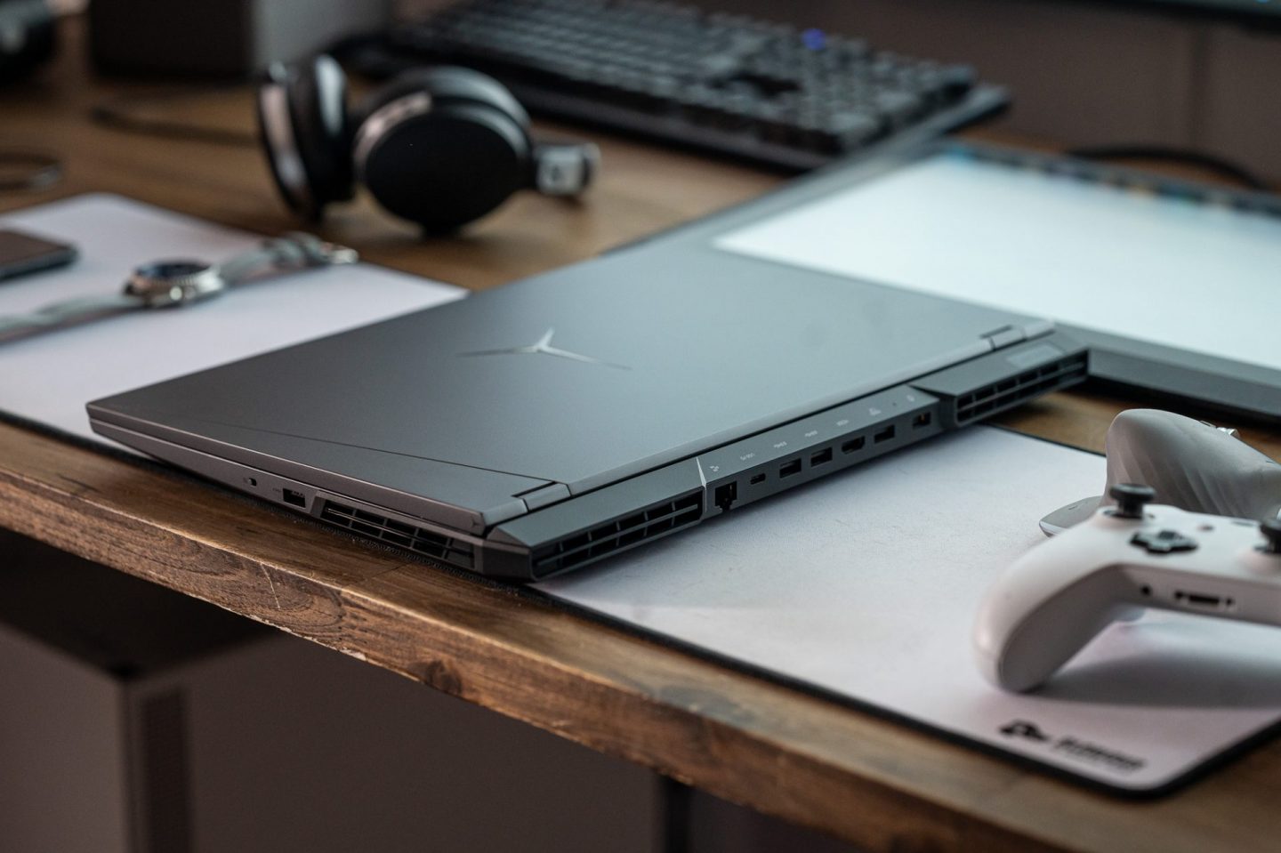 Csukott Lenovo Legion laptop az íróasztalon hátulról fotózva, jól látható csatlakozókkal, mellette kontrollerrel és fejhallgatóval.
