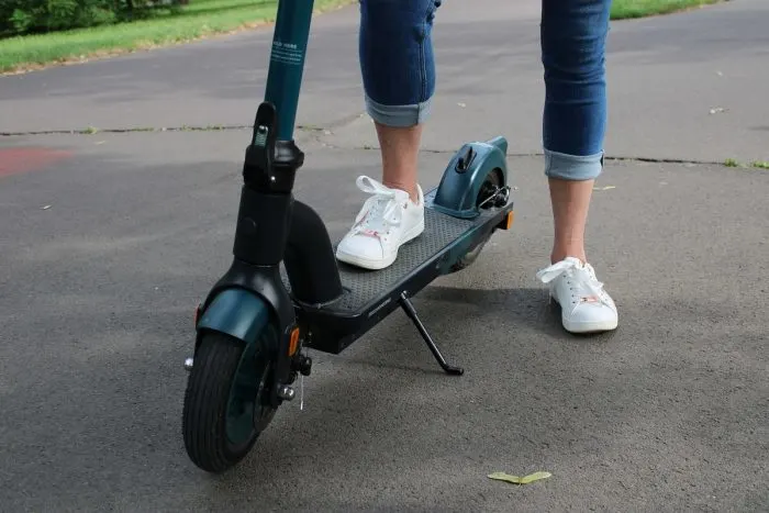 Közeli kép egy sötétkék és fekete elektromos rollerről, amin egy fehér cipőt és farmernadrágot viselő személy áll az aszfalton.