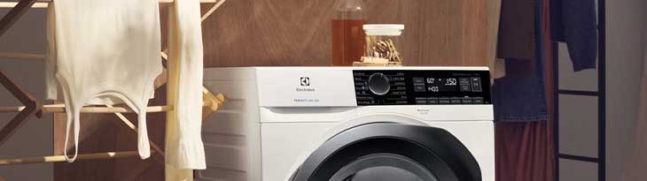 Fehér mosógép fekete ajtóval, szárítóállványon ruha, háttérben fa fal és ruhaszárító.