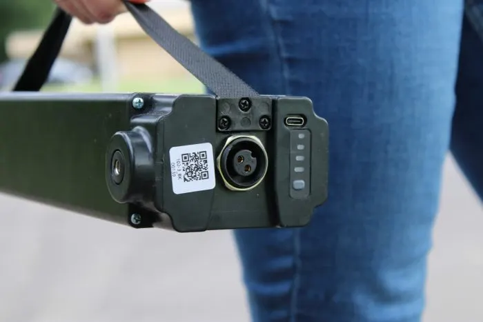 Közeli kép egy elektromos roller fekete akkumulátoráról, rajta csatlakozóval, töltöttségjelző LED-ekkel és QR-kóddal, egy ember kezében.
