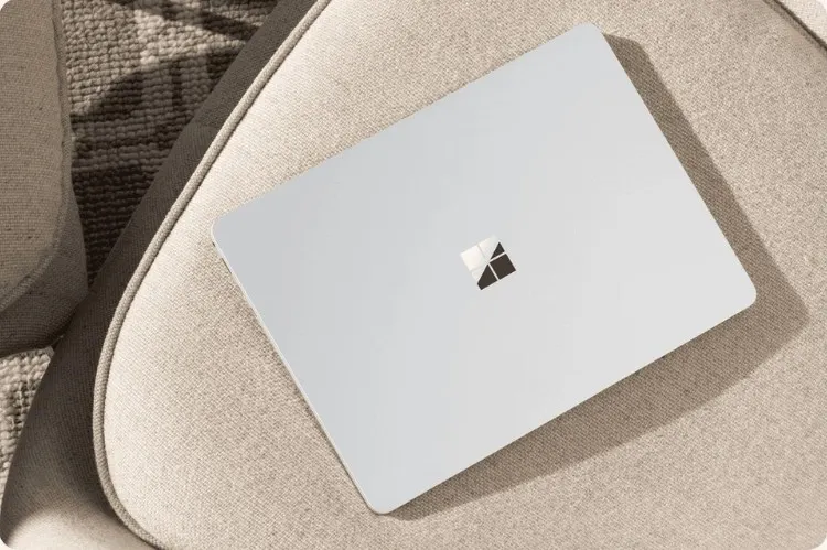 Egy szürke laptop látható egy bézs színű karosszéken, a háttérben pedig egy texturált szőnyeg.