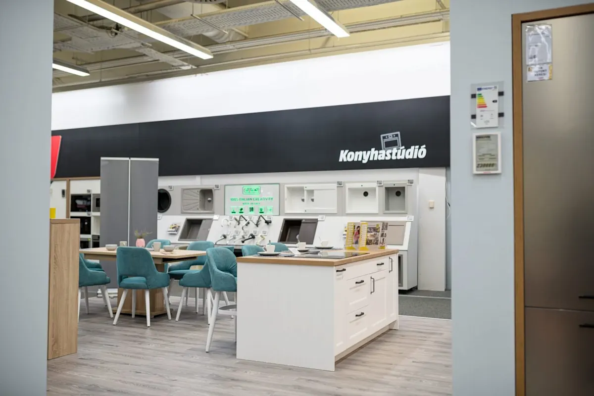 Modern konyhastúdió bemutatóterme kiállított konyhaszigettel, étkezőasztallal, türkizkék székekkel és a falon Konyhastúdió felirattal.