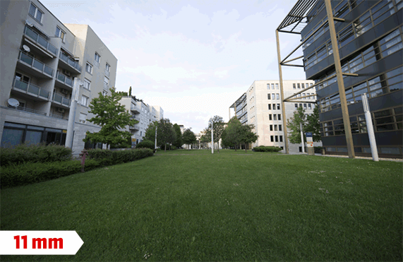 Zöld gyepes területet szegélyeznek épületek és fák, 11 mm felirat. A háttérben égbolt látható.
