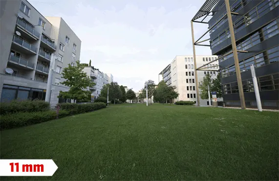 Zöld gyepes területet szegélyeznek épületek és fák, 11 mm felirat. A háttérben égbolt látható.