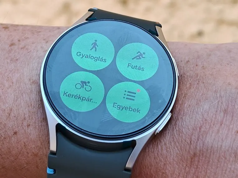 Közeli fotó egy csuklón lévő Samsung Galaxy Watch okosóráról, amelynek kijelzőjén négy zöld ikon látható a különböző edzéstípusok (gyaloglás, futás, kerékpár és egyebek) indításához.