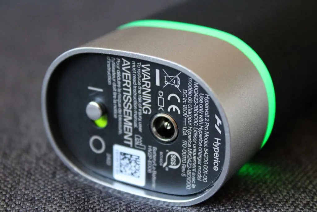 Egy Hyperice Hypervolt 2 Pro masszázspisztoly markolatának alja a technikai adatokkal, a töltőcsatlakozóval és egy világító zöld LED-gyűrűvel.