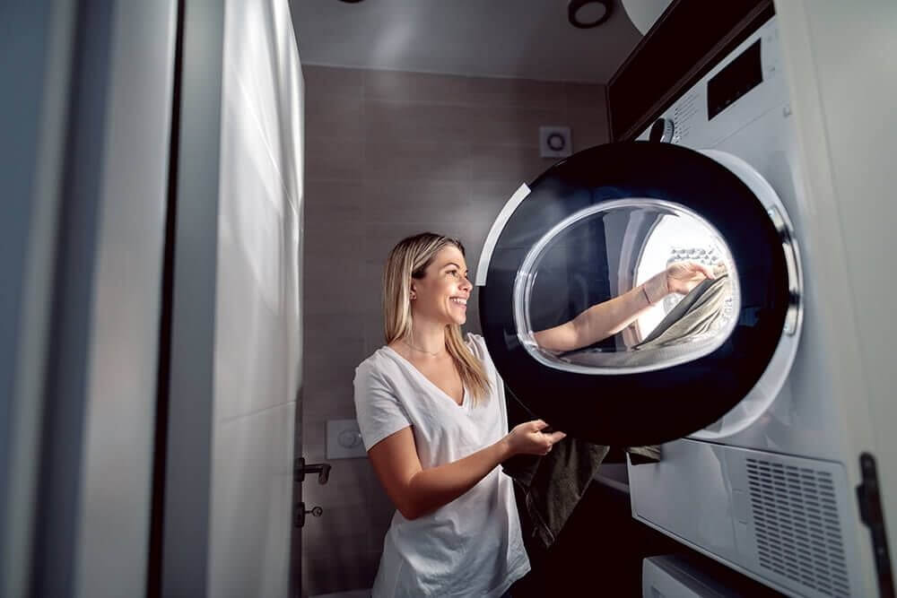 Egy nő mosógépet pakol a fürdőszobában. A gép fehér, a nő fehér pólót visel. A háttérben szürke fal és zuhanyfüggöny látható.