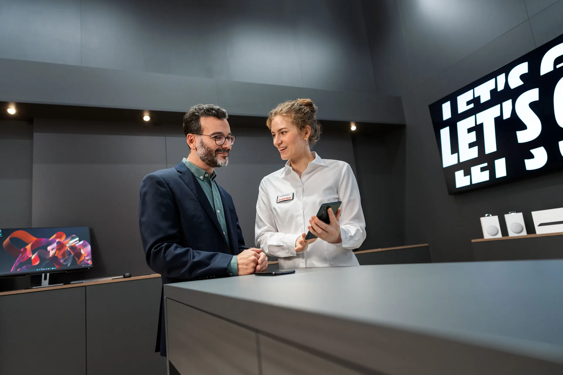 Ügyfél és eladó beszélget egy pultnál, ahol telefon és reklám látható.