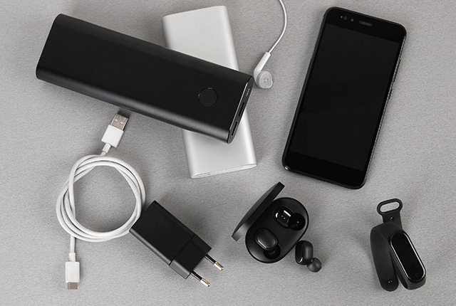 A képen egy fekete mobiltelefon, fekete és ezüst színű külső akkumulátorok, töltő, vezeték, fülhallgató és egy fekete karpánt látható szürke háttéren.