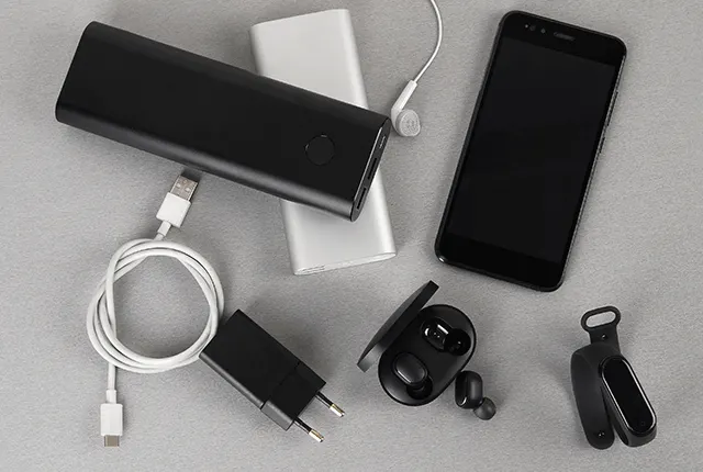 A képen egy fekete mobiltelefon, fekete és ezüst színű külső akkumulátorok, töltő, vezeték, fülhallgató és egy fekete karpánt látható szürke háttéren.