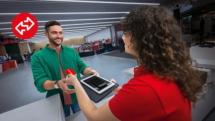 Egy férfi zöld ingben piros ajándékkártyát ad át egy táblagépet tartó piros ruhás személynek, üzlethelyiségben.