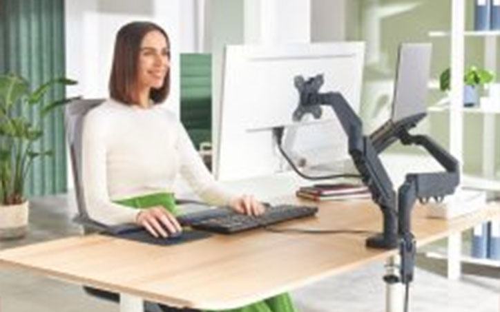 Nő ergonomikus irodai munkaállomásnál dolgozik monitor- és laptopkarral felszerelt íróasztalnál.