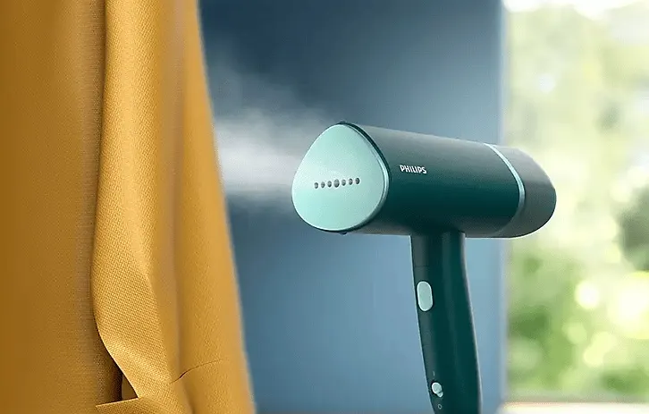 Philips kézi ruhagőzölő működés közben, amint gőzt bocsát ki egy sárga függöny mellett.