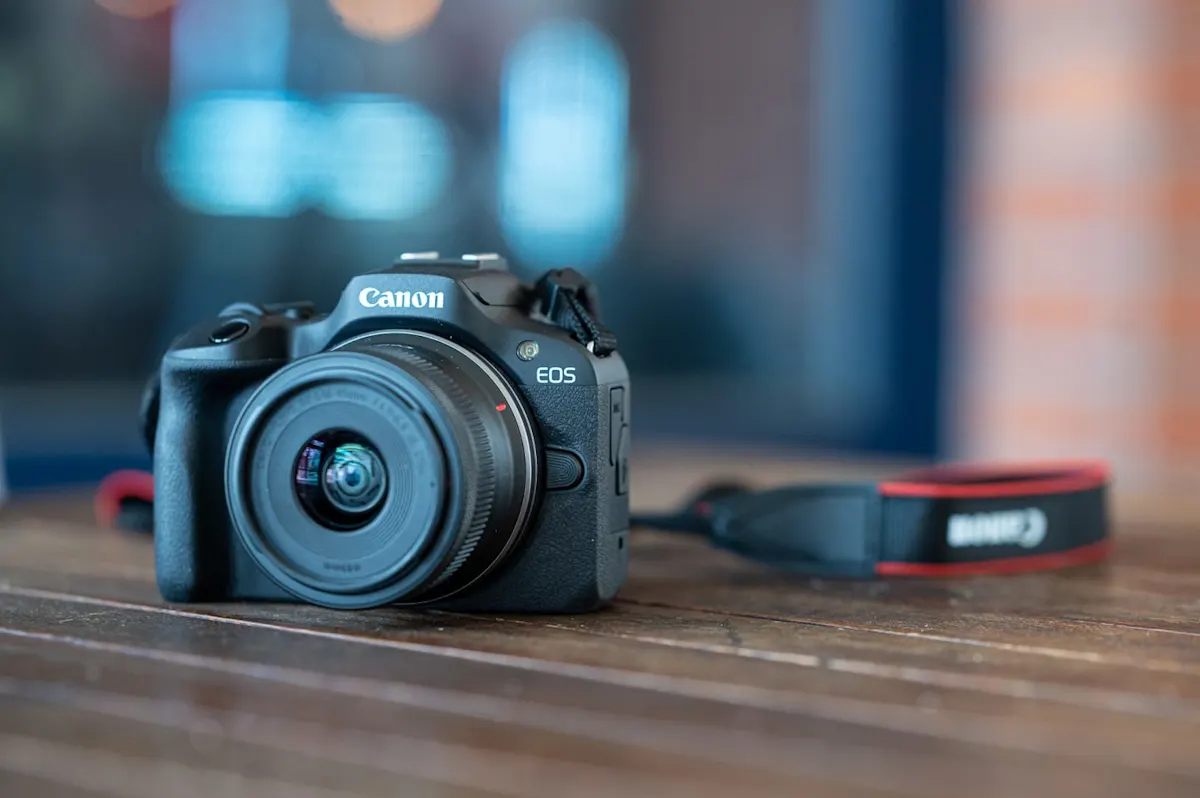 Fekete Canon EOS fényképezőgép objektívvel egy barna faasztalon, mellette egy piros-fekete Canon nyakpánttal, elmosódott háttér előtt.