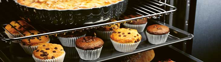 Sütőben muffinok papírban, csokoládé darabokkal és egy nagy sütemény fekete tálban, rácson.