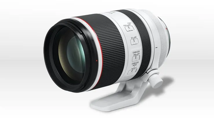 Egy fehér és fekete színű, piros csíkkal díszített Canon telefotó objektív (L-szériás), állványgyűrűvel felszerelve, fekvő pozícióban.