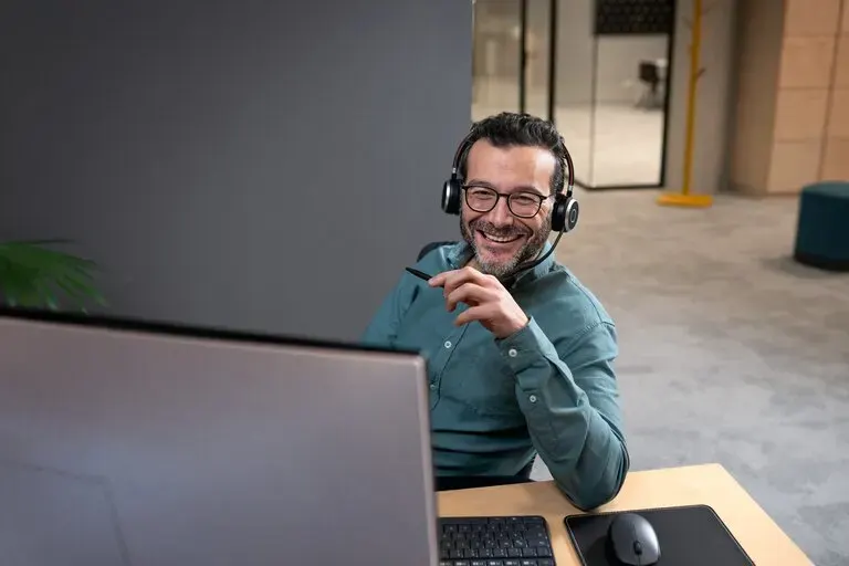 Mosolygó férfi fejhallgatóval, szemüveggel, asztalnál ülve, számítógép előtt, kezében tollal.