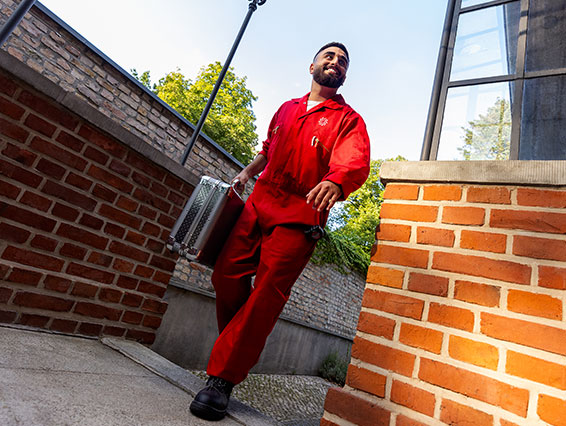 Férfi piros munkaruhában sétál, fém bőröndöt visz. Háttérben téglafal, fa és épületablak látható.