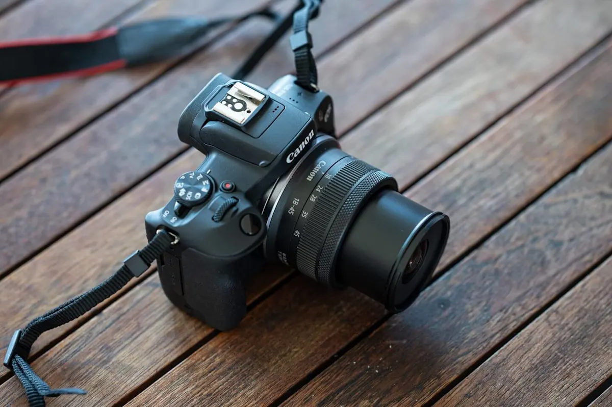 Fekete Canon tükör nélküli fényképezőgép felülnézetből, kitolt 18-45mm-es objektívvel és vállpánttal egy barna fapadlón elhelyezve.